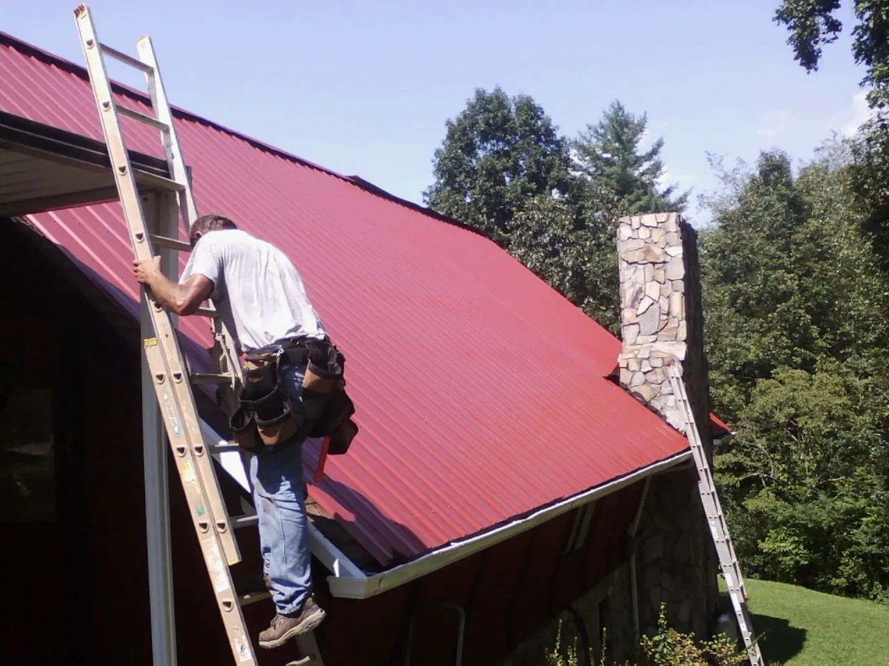 Roofer on a ladder beside a red metal roof and stone chimney — Soffit & Fascia Repair by Roofing Brothers in Winnabow