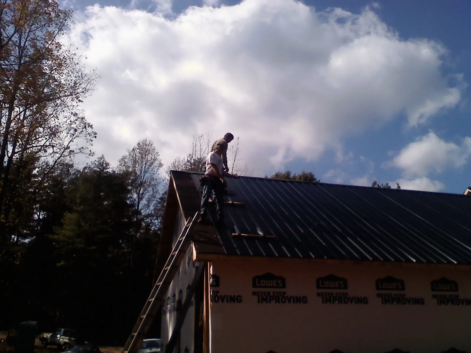 Roofer fastening dark metal panels high against a cloudy sky, from a Roof Inspection job in Winnabow